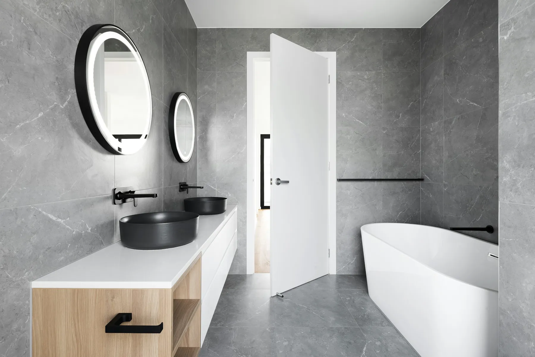 A stone-clad bathroom with a floating oak vanity, black fixtures and a freestanding tub